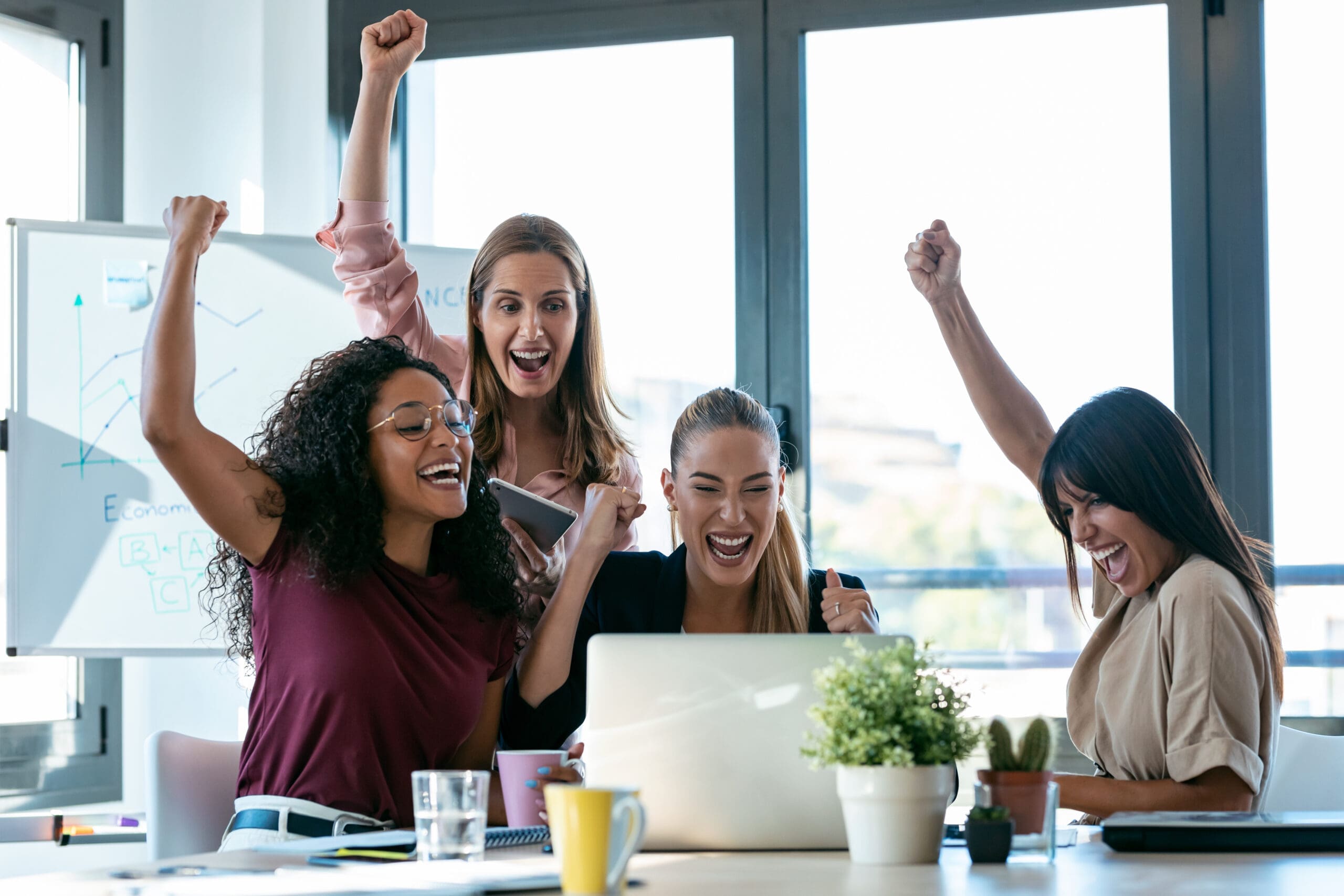 Four happy smart business women work with laptops on the desk while celebrating a victory in the coworking space in the office.